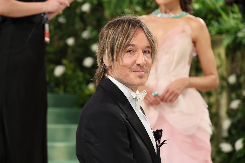 Keith Urban in a tuxedo with a bow tie on a red carpet event, with someone in a formal, ruffled gown in the background