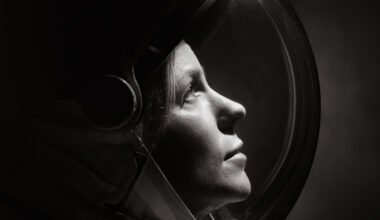 A black and white photo of a woman wearing a spacesuit and helmet. She looks upward toward the right. The photo shows only her head and shoulders.