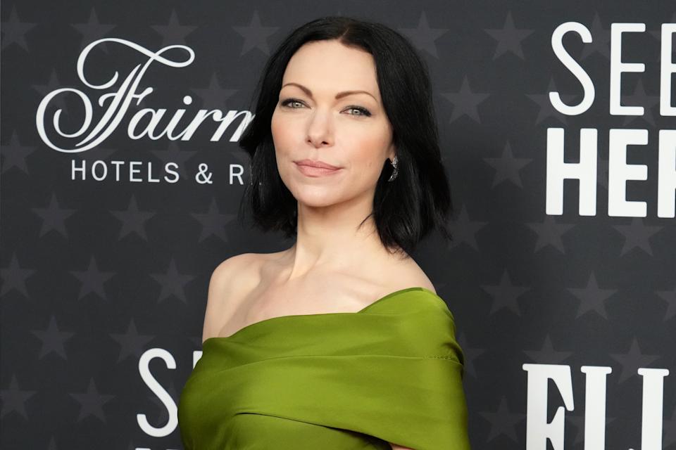 Laura Prepon on a red carpet wearing an elegant off-the-shoulder gown with sleek, straight hair