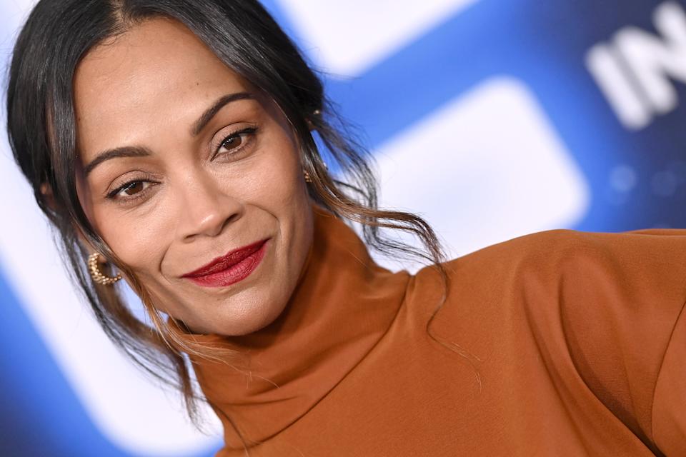 Zoe Saldana in a high-neck top poses at a press event, smiling with her hair styled back