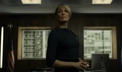 A person stands confidently in an office with a U.S. flag and a computer in the background