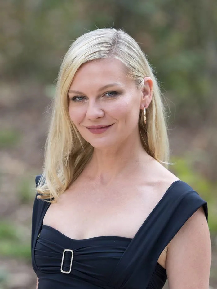 Kirsten with long hair in an elegant, sleeveless dress, smiling at an outdoor event