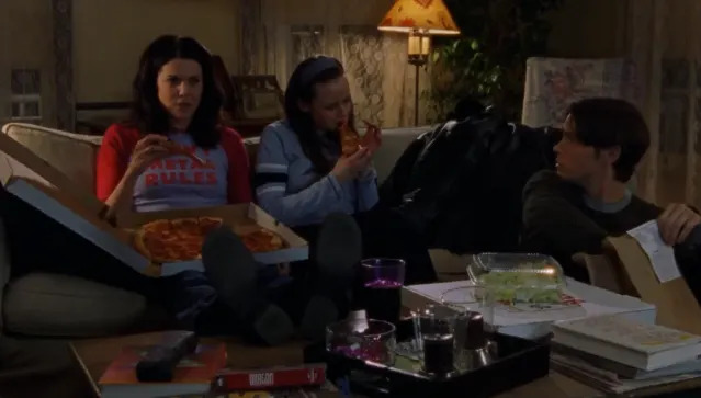 Three people sit on a couch eating pizza. One holds a pizza box while another eats a slice. They are engaged in casual conversation