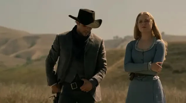 A man in a cowboy hat and a woman in a long dress stand outdoors in a grassy landscape, reminiscent of a Western scene
