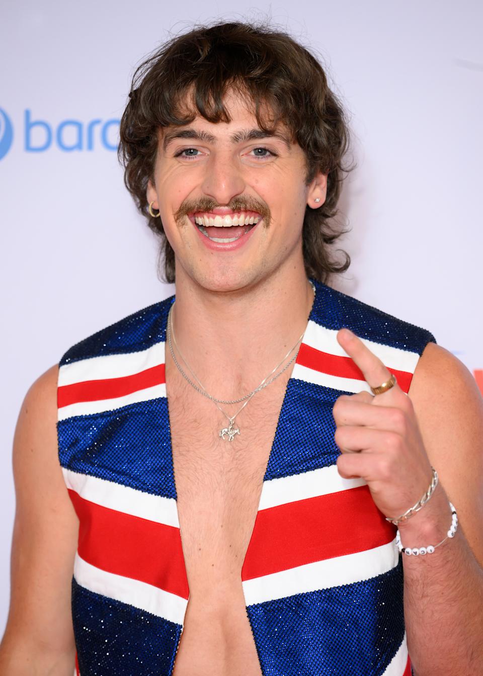 Benson Boone smiling, wearing a sleeveless, patterned outfit with necklaces, poses on a red carpet