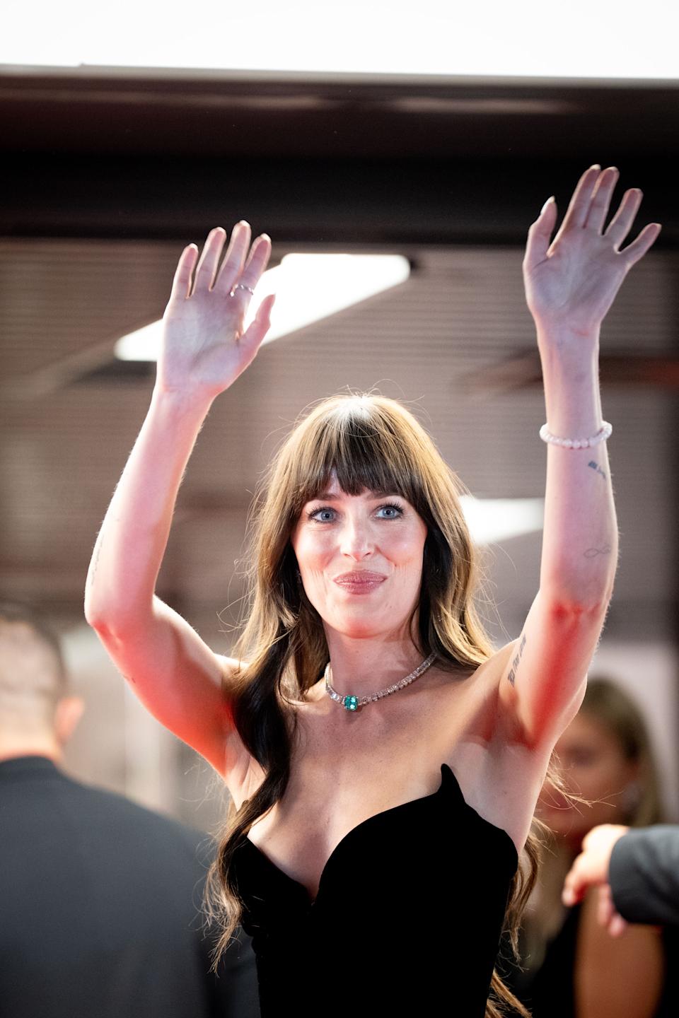 Dakota Johnson on the red carpet, waving, in a strapless, fitted gown with long hair and a necklace, smiling at the camera