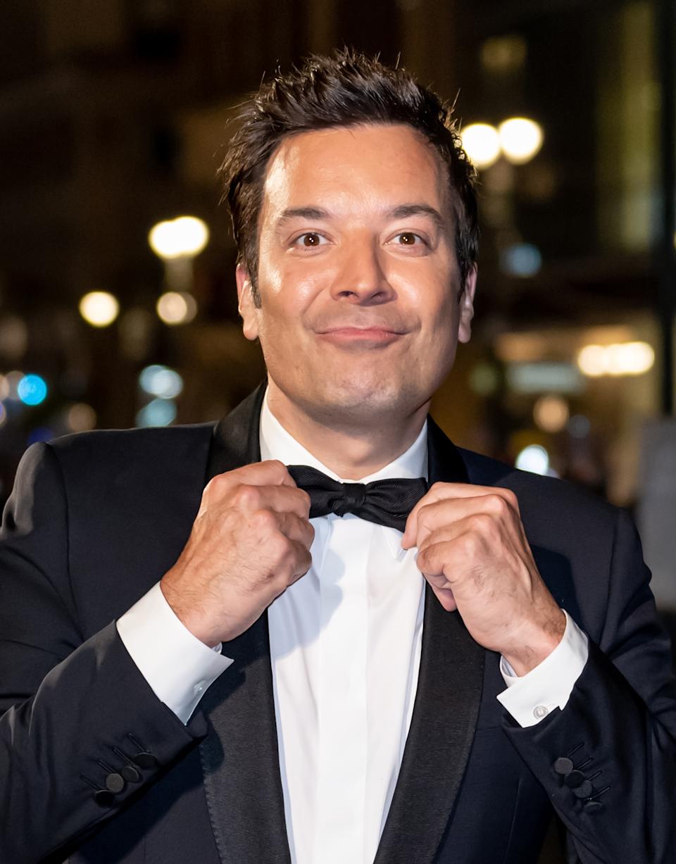 Jimmy Fallon in a tuxedo adjusts a bow tie while smiling, standing in a well-lit environment