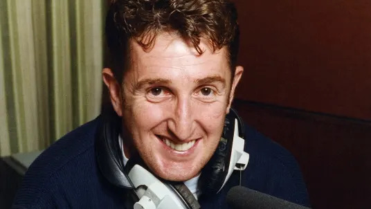 A man with curly brown haior wearing a navy fleece and smiling at the camera. He has headphones around his neck and is sat in front of a black microphone. It is a head and shoulders shot of him. 