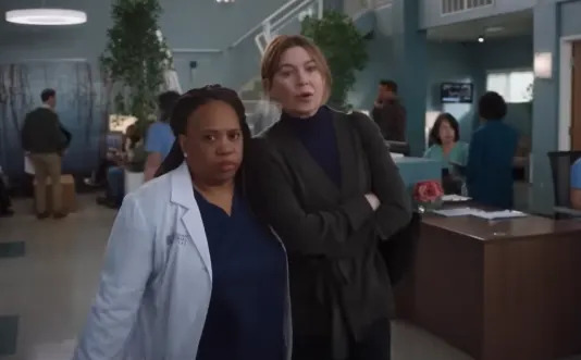 Two women in a modern hospital setting. One wears a lab coat over scrubs, the other a dark cardigan. Staff and patients are in the background