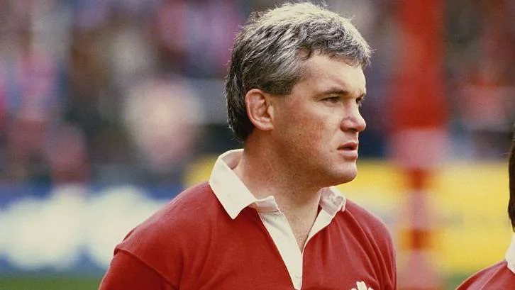 A man with grey-brown hair wearing a red Welsh rugby jersey. He has a serious expression on his face as he looks off to the side. There's a blurry background of a rugby stadium behind him. 