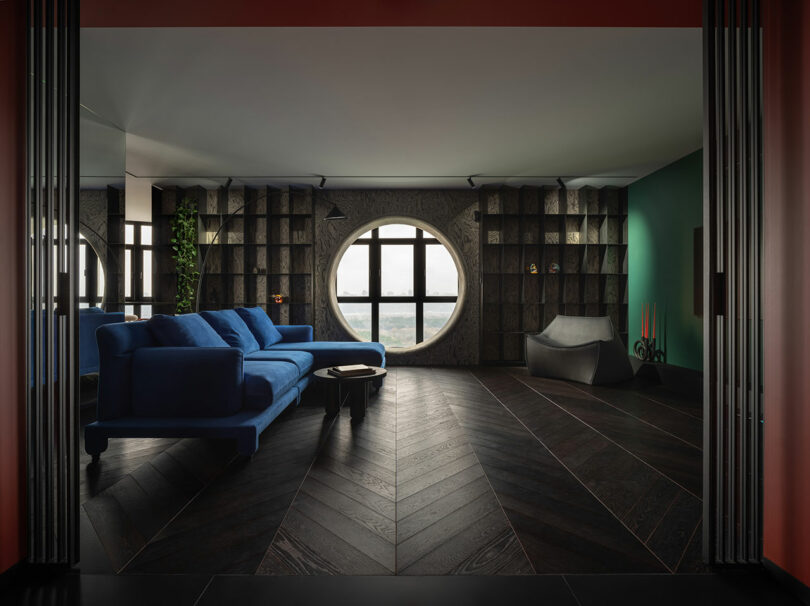 Modern living room with a blue sofa, circular window, dark wood floors, built-in shelving, and green accent wall. Sculptural gray chair and candles add decorative touches.