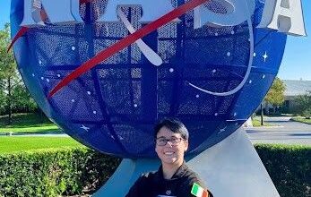 A person wearing a black NASA flight suit stands smiling with arms crossed in front of the large blue NASA globe sculpture. The Irish flag patch is visible on their sleeve, and the sky is clear and blue in the background.