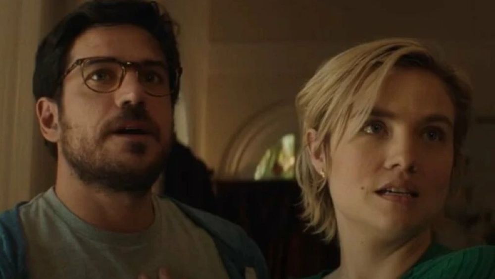 Diego and Sage, a Brazilian man and a white woman, stand in a dimly lit mansion while looking at something out of frame in shock in a still from the horror movie Bone Lake.