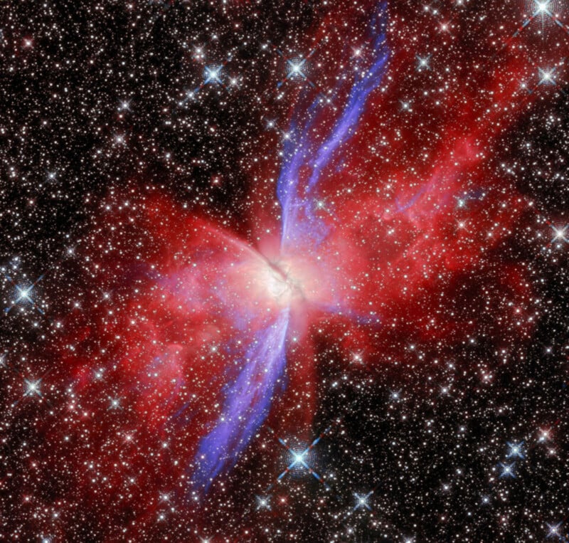 A bright galaxy at the center emits glowing red and blue gas plumes, surrounded by countless stars against a dark sky. The colorful jets extend outward in X-shaped formations.