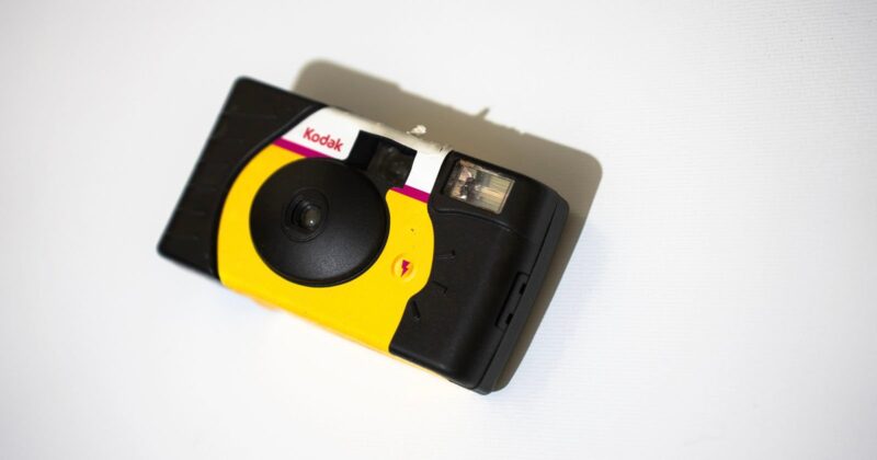 A Kodak disposable film camera with a yellow and black body and red accents, placed on a white surface, viewed from above.