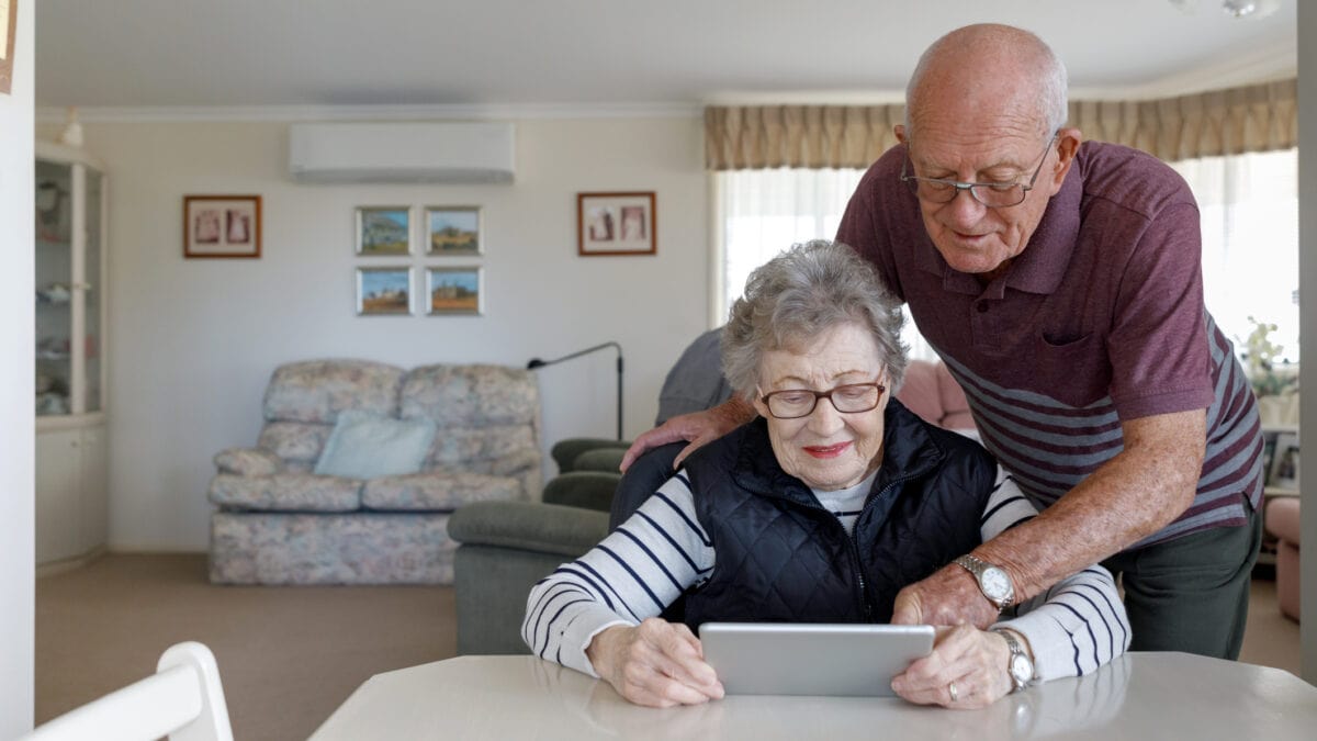 An older gentleman leans over his partner's shoulder as she looks at a tablet device while seated at a table.