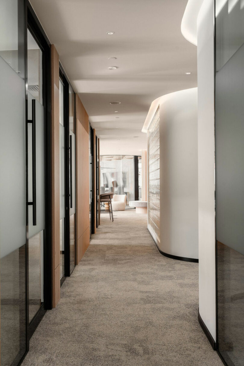 Modern office corridor with glass-walled rooms on the left, curved wall on the right, carpeted floor, and a seating area visible at the end near large windows.