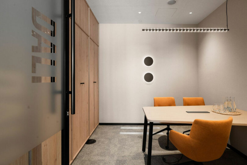 Small modern meeting room with a wooden table, two orange chairs, wall-mounted shelves, round wall accents, and a glass door labeled "DALI." Three glass water bottles sit on the table.