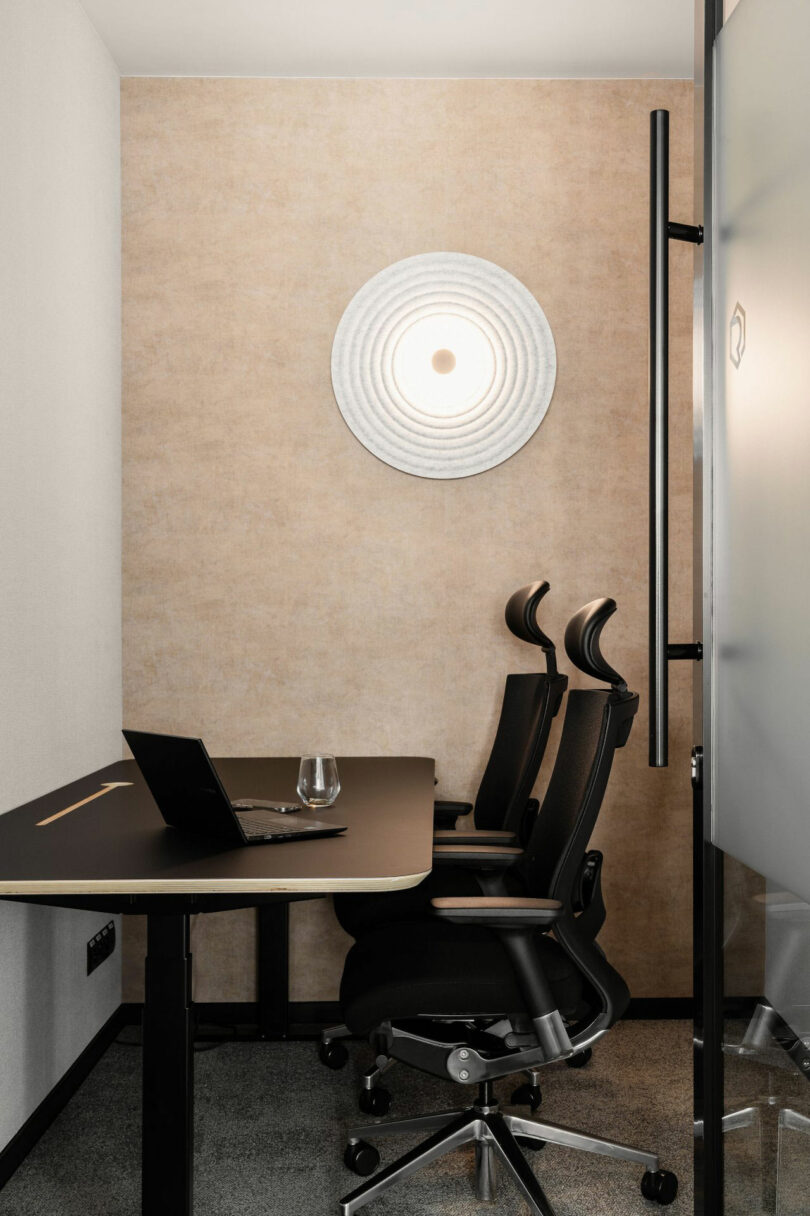 Small modern office with two black ergonomic chairs, a black desk, an open laptop, a glass of water, and a circular wall light on a beige wall.