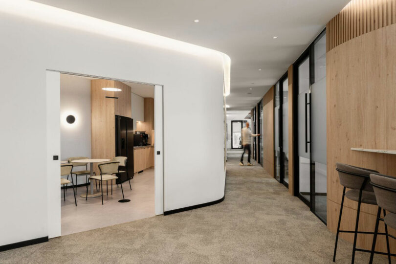 Modern office corridor with beige carpet, glass-walled offices on the right, a kitchenette with tables and chairs on the left, and a person walking in the background.