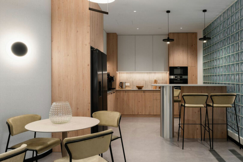 Modern kitchen with light wood cabinets, black appliances, a round table with four chairs, a bar counter with stools, and a decorative tile accent wall.
