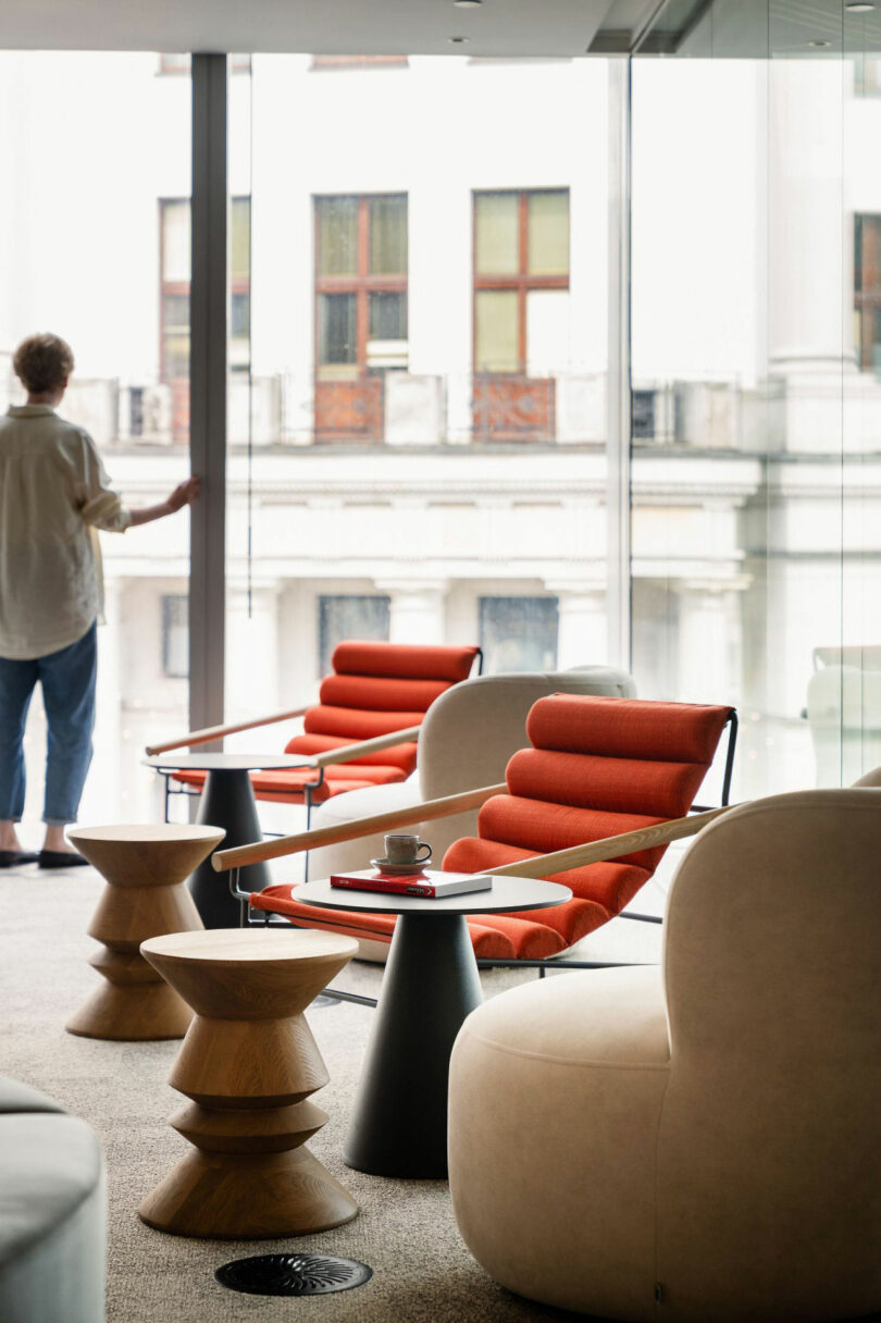 A modern lounge with red accent chairs, round tables, and a cup on a tray. A person stands by large windows overlooking a building across the street.