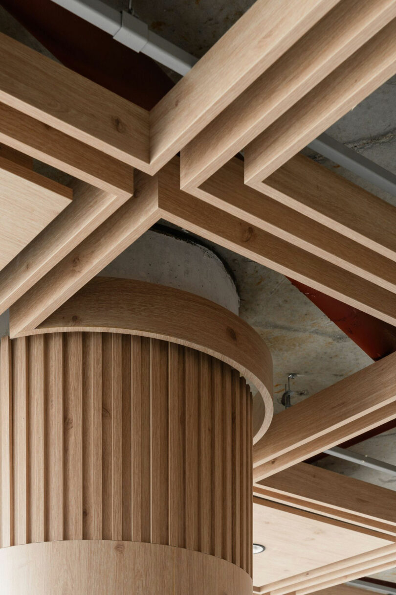 A geometric wooden ceiling design with intersecting beams and a fluted cylindrical column set against an exposed concrete and metal background.