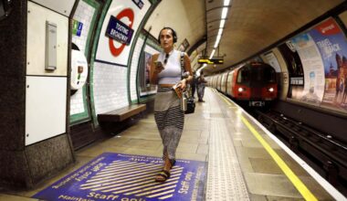 2JHRXY3 London, England, UK. London Underground - young woman with mobile phone and wearing headphone walking along the platform of Tooting Broadway station
