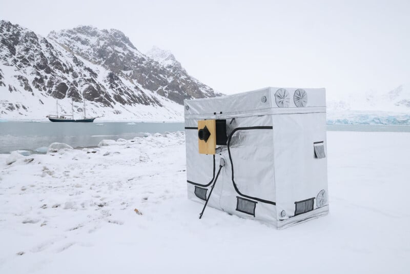 A small, box-shaped silver shelter stands on a snowy shore near icy water, with snow-covered mountains and a ship in the background. The shelter has vents and a small yellow device attached to its side.
