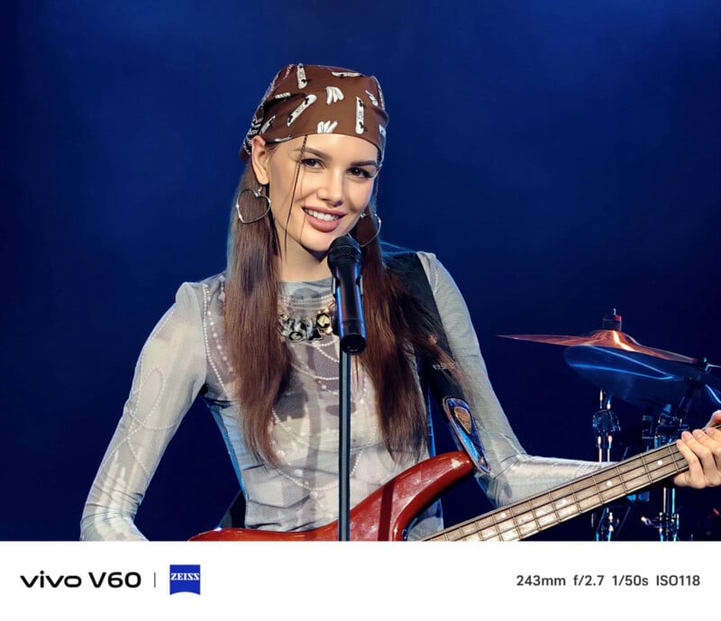 A smiling woman wearing a brown bandana and hoop earrings plays an electric guitar and sings into a microphone on stage, with blue lighting and drums visible in the background.