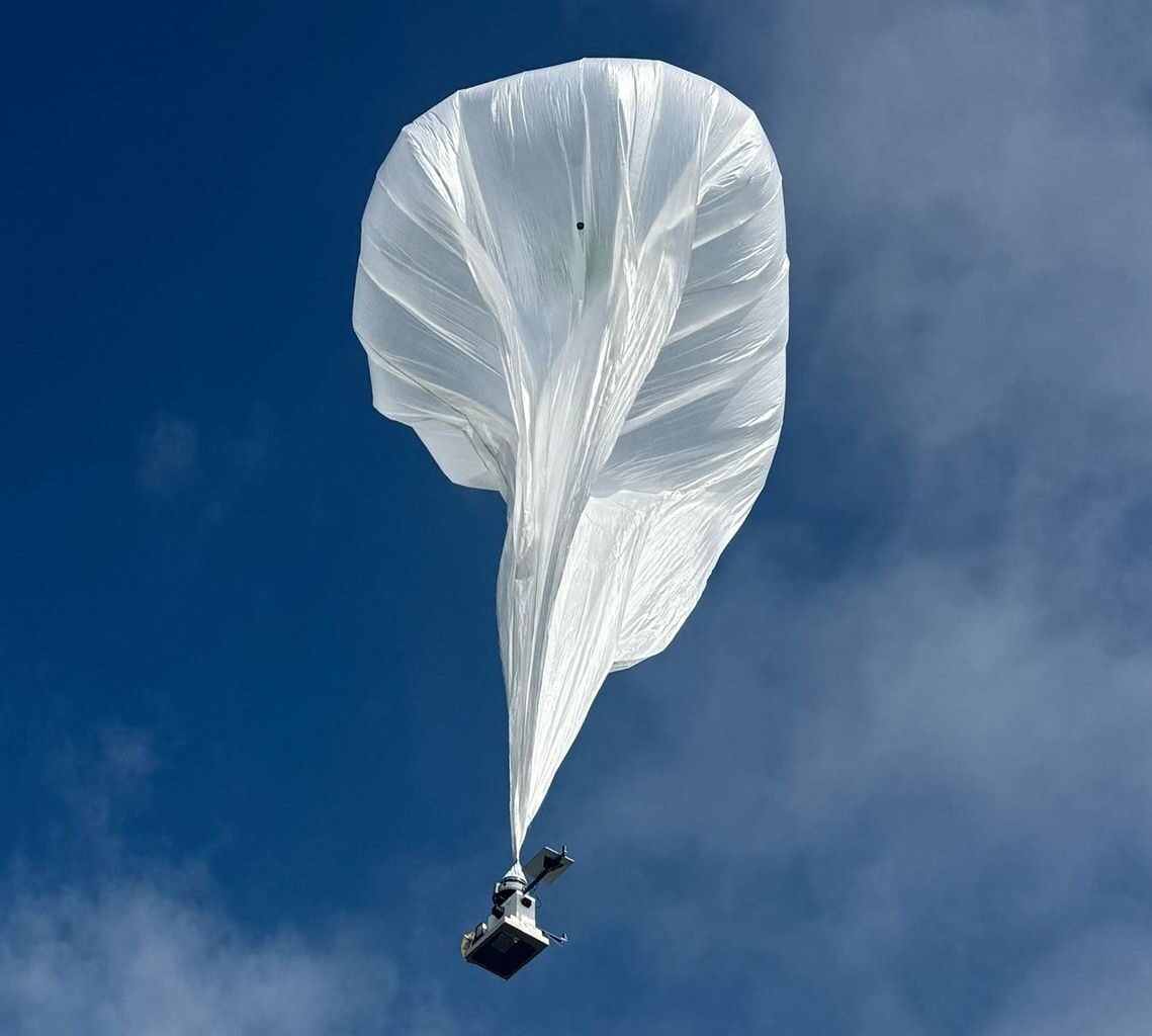 A hot-air balloon hoisting a complex radar instrument, mid-flight.