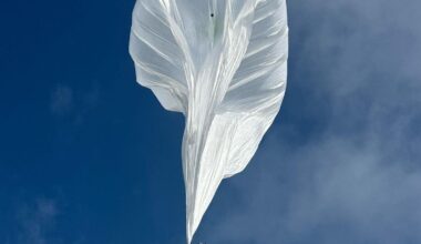 A hot-air balloon hoisting a complex radar instrument, mid-flight.
