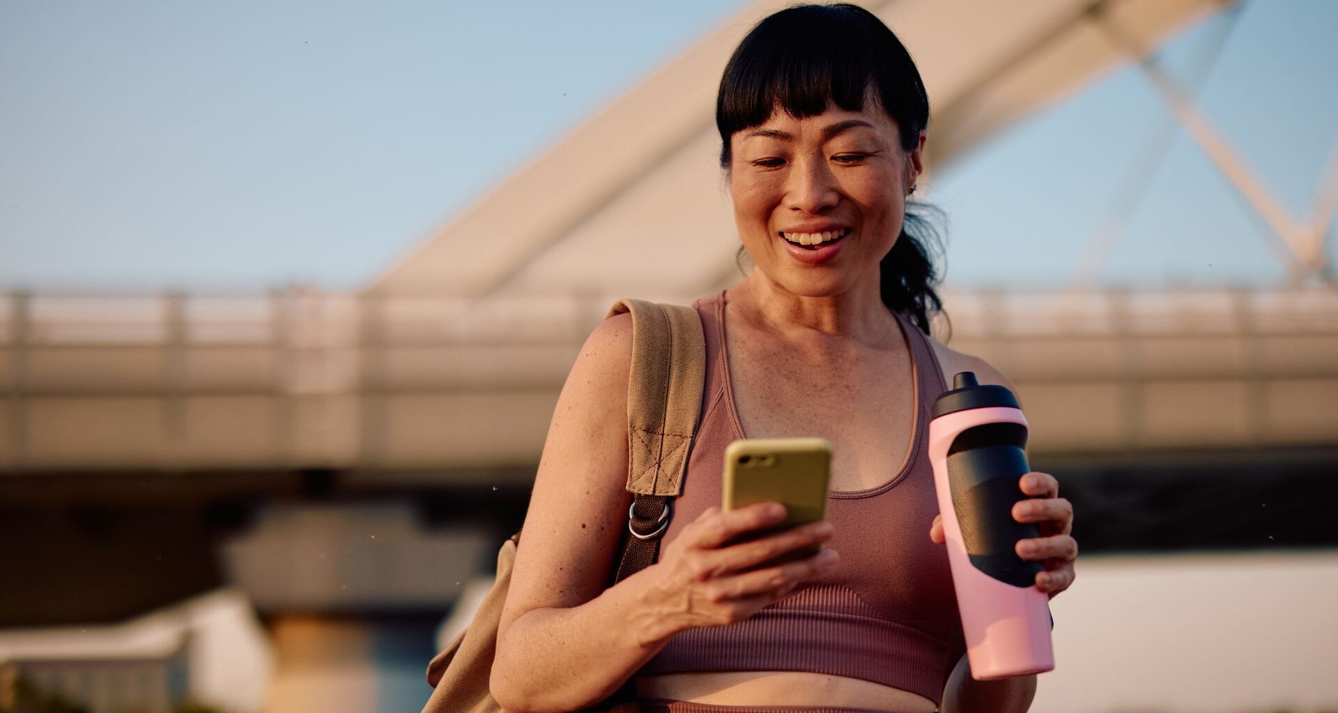 Woman holding bottles of electrolytes, smiling and looking at phone while outside in hot weather