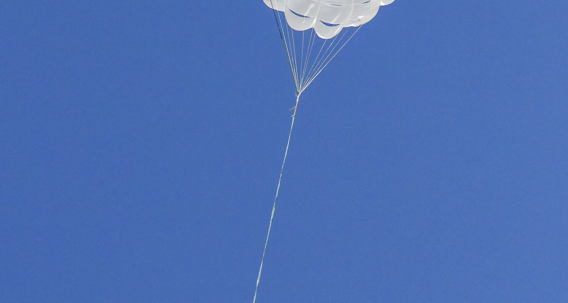 A parachute carrying a capsule is fully open against a canvas of deep blue sky.