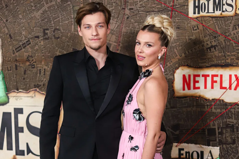 Jake Bongiovi and Millie Bobby Brown pose together on the red carpet, with Bongiovi in a black suit and Brown in a pale pink gown decorated with black floral accents.