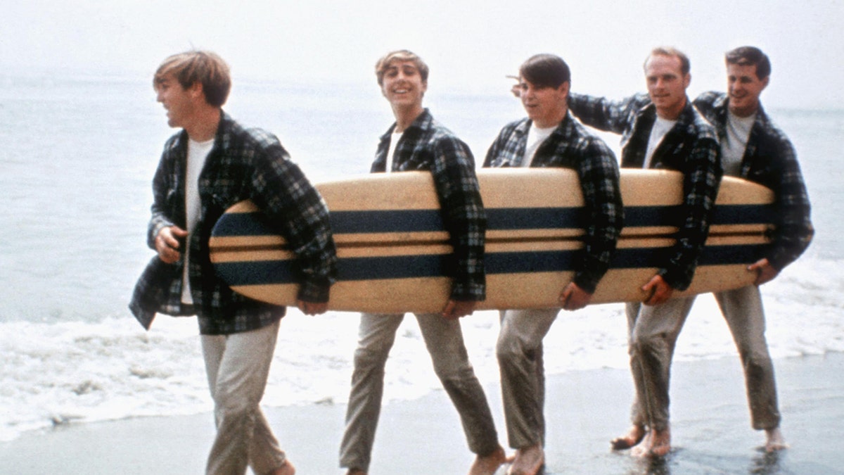 The Beach Boys walking on the beach holding a surfboard.
