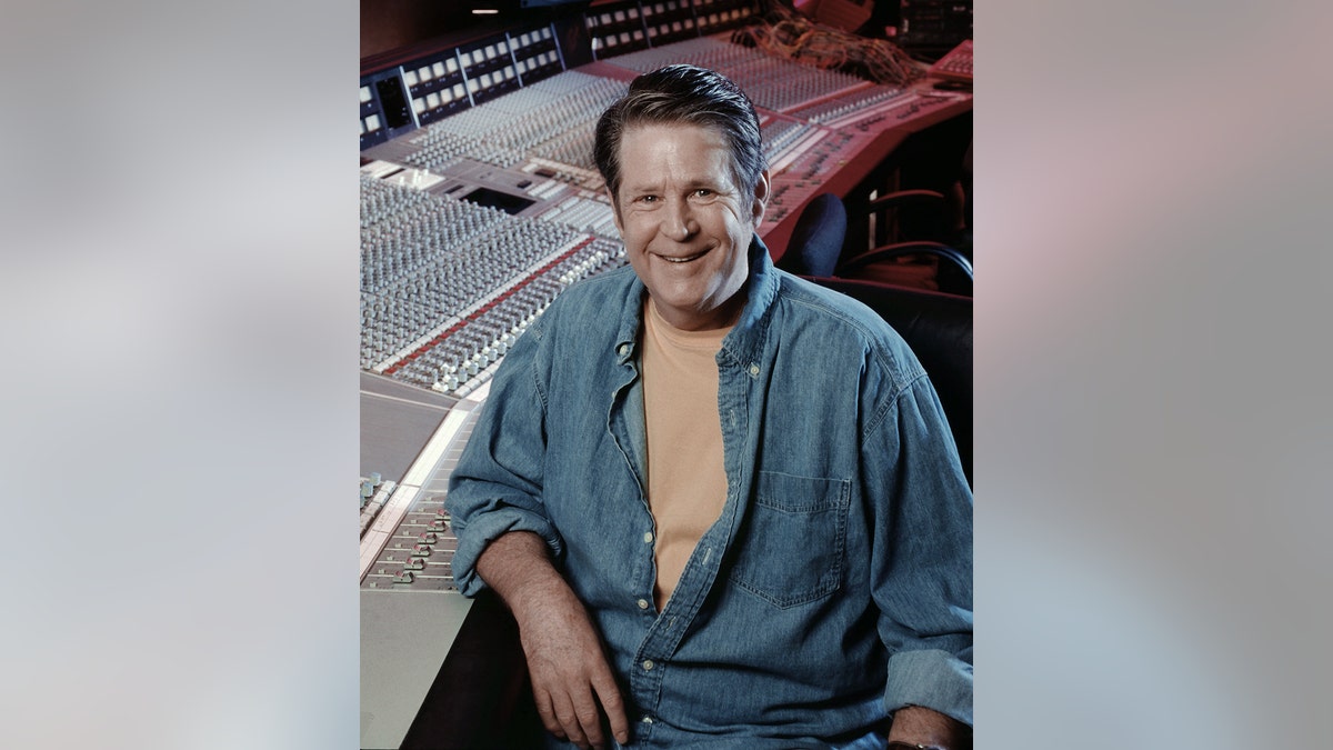 Brian Wilson smiling in front of a studio wearing a denim open shirt with a yellow t-shirt.