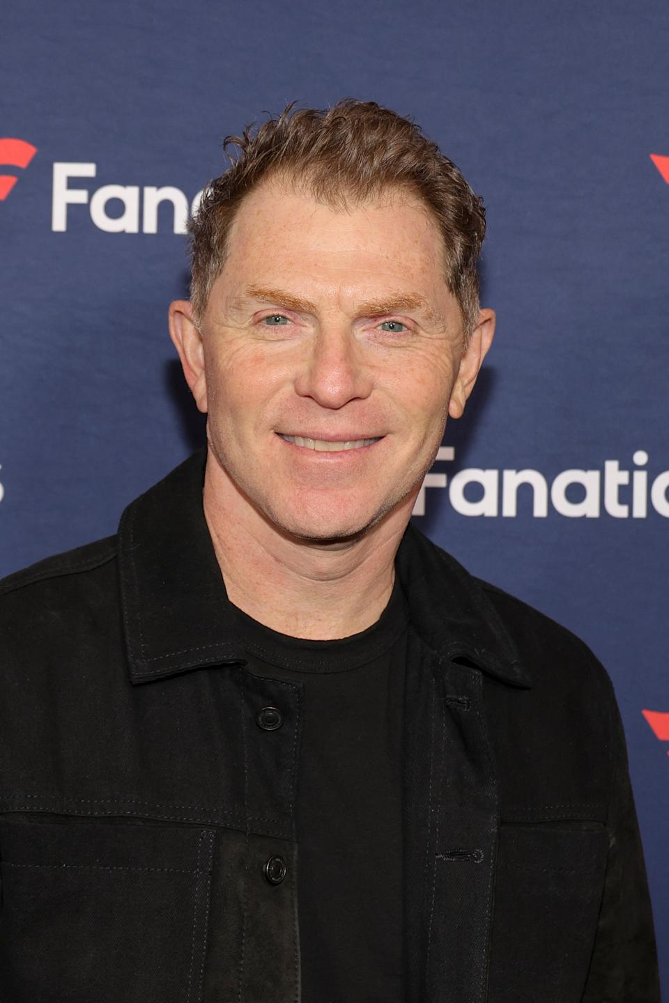 Bobby Flay at a Fanatics event, wearing a jacket and smiling, stands against a branded backdrop