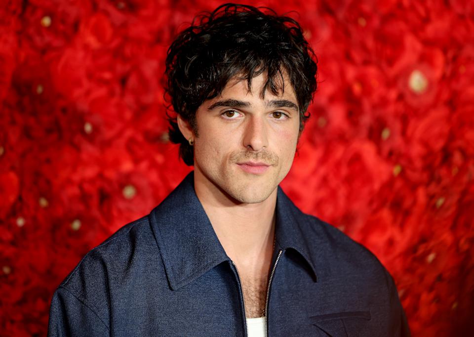 Jacob Elordi with curly hair in a casual jacket poses against a detailed floral backdrop