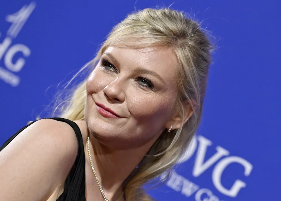 Kirsten Dunst in elegant attire poses confidently at an event, against a backdrop with a repeating logo
