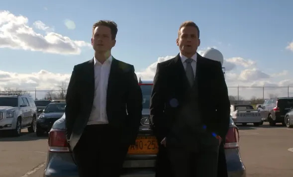 Two suited men leaning against a parked car in a parking lot, with a cloudy sky in the background