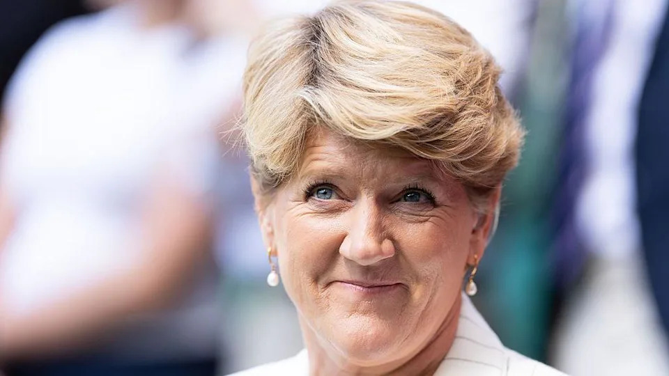 A woman with short blonde hair wearing a white jacket and gold pearl-drop earrings. She is looking off to the side and smiling. It is a headshot. 