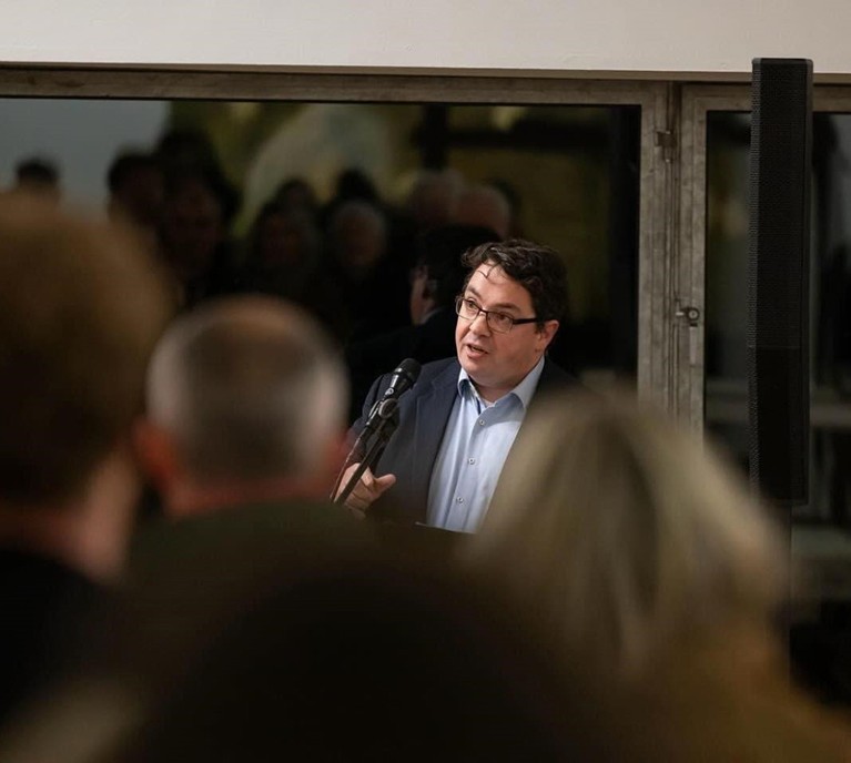 A man in a suit talks to an out of focus crowded room.