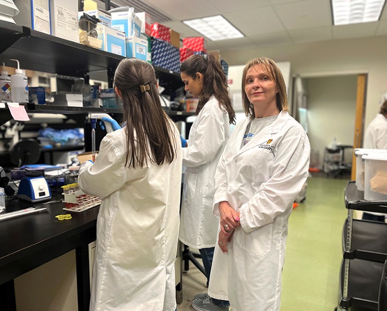 A female doctor in a busy lab looks at the camera. Two figures work on lab stools behind her.