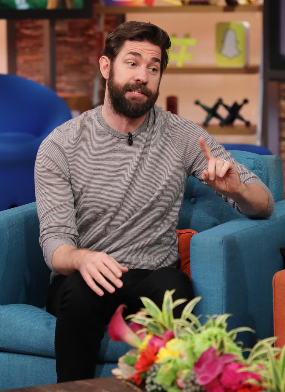 John Krasinski in a casual striped shirt gestures with his hand while sitting on a sofa during an interview