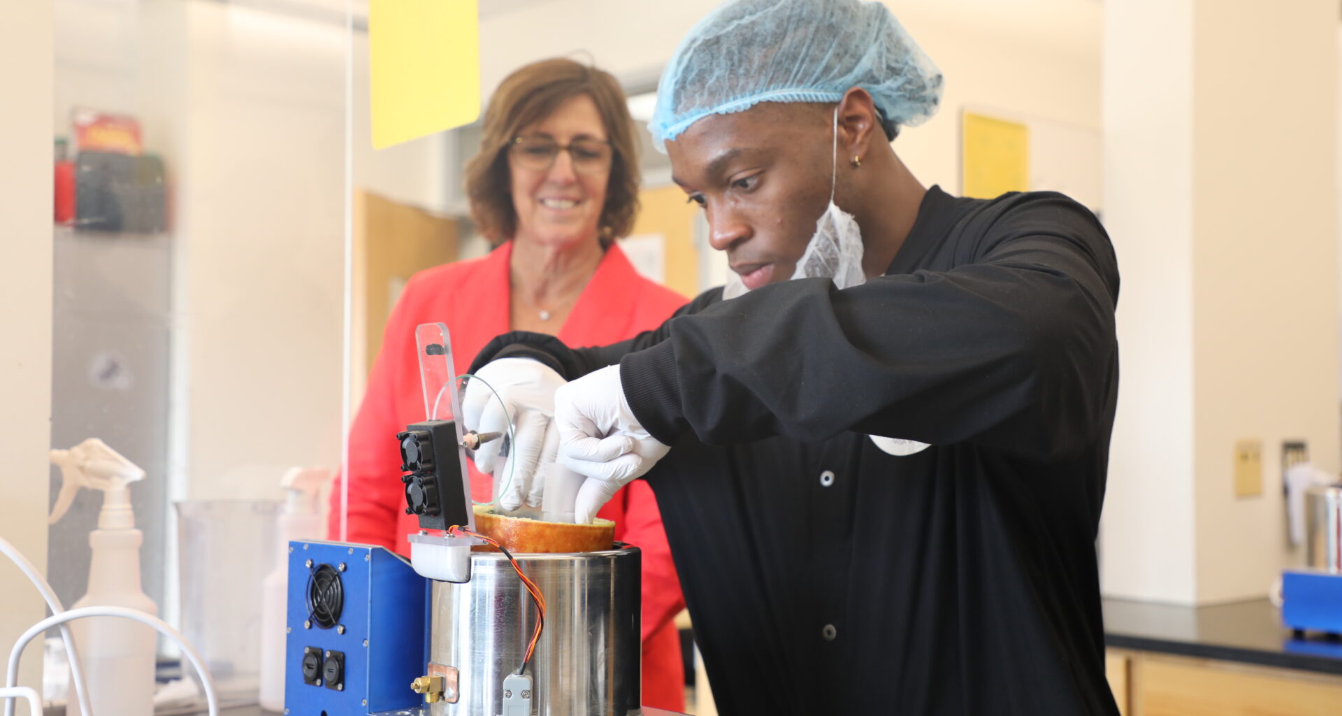 A man wearing gloves and a hairnet uses a flat spatula-like mechanism to remove a cylindrical omelet from a small cylindrical oven, called SATED. Behind him, a smiling woman observes the food.
