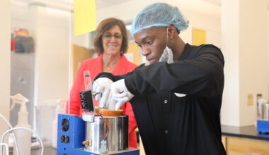 A man wearing gloves and a hairnet uses a flat spatula-like mechanism to remove a cylindrical omelet from a small cylindrical oven, called SATED. Behind him, a smiling woman observes the food.