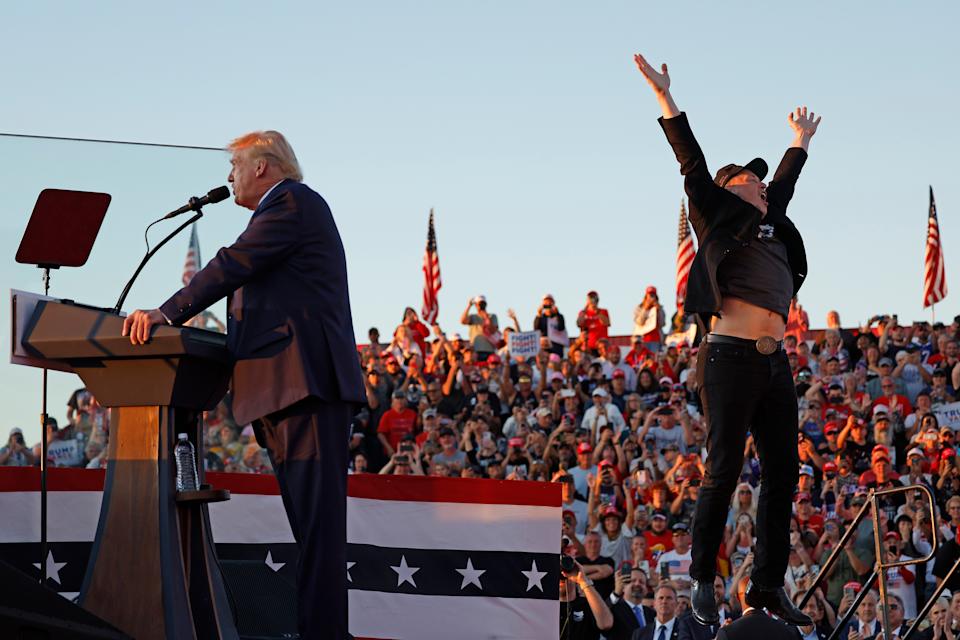 Donald Trump stands at a podium giving a speech before a large crowd as Elon Musk jumps onstage