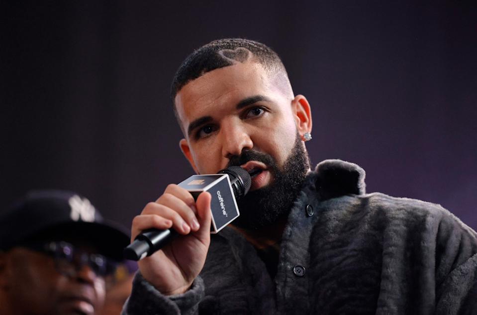 Drake speaks into a microphone at an event, wearing a textured jacket