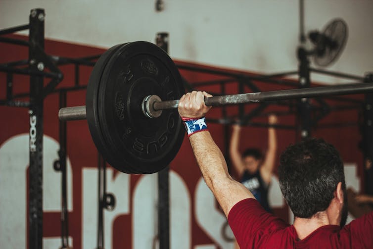 A man in a red T-shirt seen from behind, lifting a barbell over his head